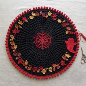 May include: A circular black crochet doily featuring a central red spiderweb motif, surrounded by a wreath of crocheted autumn leaves and acorns in shades of red, orange, and yellow, finished with a red pom-pom border and a small red ghost.