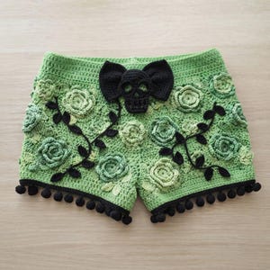 May include: Light green crochet shorts featuring a black skull and bow applique at the center waist. The garment is decorated with various shades of green crocheted floral motifs, black vine accents, and a black pom-pom trim along the bottom hem.