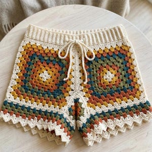 May include: Crochet shorts featuring a granny square pattern in shades of mustard yellow, forest green, and burnt orange. The shorts have an off-white ribbed waistband with a matching drawstring tie and a scalloped edge along the bottom hem.