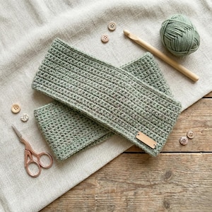 May include: A handmade, sage green crochet headband and matching wrist cuffs. A ball of yarn, wooden crochet hook, small scissors, and wooden buttons are also visible. The headband has a small, blank, rectangular tag.