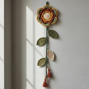 Puede incluir: Un colgante de pared de flores de ganchillo hecho a mano. La flor tiene capas de pétalos naranjas, amarillos y verdes. Un tallo verde con hojas verdes y beige se extiende hacia abajo, con borlas y cuentas de madera en la parte inferior. Para colgar en la pared.