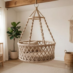 May include: A beige macrame baby swing suspended from the ceiling. The swing features a round, woven base with a diamond pattern and a small pillow. It is supported by knotted rope and a wooden ring.