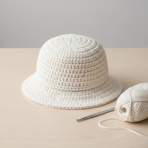 Peut inclure: Un chapeau seau en crochet de couleur crème avec une couronne arrondie et un petit bord. Une pelote de laine crème et un crochet sont à côté du chapeau. Le chapeau et la laine sont sur une surface en bois clair.