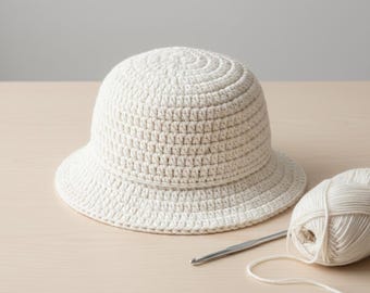 May include: A cream-coloured, crocheted bucket hat with a rounded crown and a small brim. A ball of cream-coloured yarn and a crochet hook are next to the hat. The hat and yarn are on a light-coloured wooden surface.
