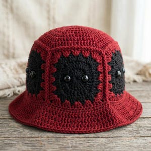Op de afbeelding: Een handgemaakte gehaakte emmerhoed in rood en zwart. De hoed heeft een rode kroon en rand, met zwarte, bloemvormige accenten aan de zijkanten. Kleine, zwarte kralen zijn op de zwarte delen genaaid. De hoed wordt gepresenteerd op een houten oppervlak.