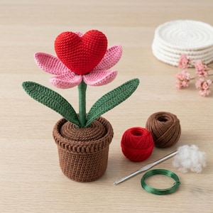 Könnte beinhalten: Handgefertigte Häkelblume mit roter herzförmiger Blüte, rosa Blütenblättern und grünen Blättern, in einem braunen Häkeltopf. Zusätzliche Bastelmaterialien sind enthalten.