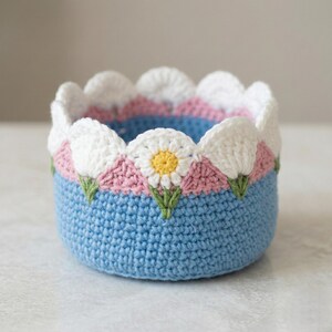 Op de afbeelding: Een ronde, gehaakte mand in lichtblauw, versierd met een geschulpte rand. De rand is versierd met witte, roze en groene bloemmotieven. De mand is waarschijnlijk gemaakt van zacht garen en is geschikt voor het opbergen van kleine items.