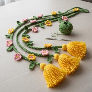 Op de afbeelding: Gehaakte bloemenslinger met gele kwasten. De slinger heeft groene wijnranken met roze en gele bloemen en groene bladeren. Een bol groen garen en een haaknaald zijn ook zichtbaar.