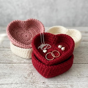 Könnte beinhalten: Gehäkelte herzförmige Schmuckschalen in Rot, Rosa und Creme. Die rote Schale enthält eine silberne Halskette mit Herzanhänger, zwei goldene Ringe und Perlenohrringe. Die anderen Schalen sind leer.