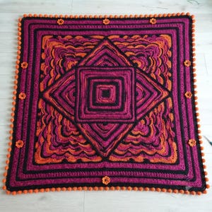 May include: A square crochet blanket featuring a geometric pattern in shades of magenta, black, and orange. The design includes a central diamond motif, concentric square borders, and a scalloped edge finished with small orange pom-poms and flowers.