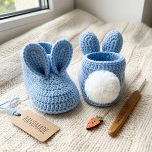 May include: Two light blue crochet bunny-themed items. One is a slipper with bunny ears, the other is a container with a white pom-pom tail. A wooden carrot button and a crochet hook are also present. A tag reads "HANDMADE".