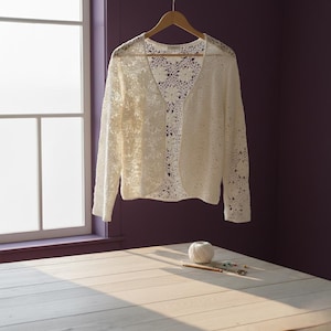 Puede incluir: Un cárdigan crema con frente abierto y un delicado diseño de ganchillo calado cuelga de una percha de madera. La prenda de manga larga se exhibe contra una pared púrpura cerca de una ventana. Una madeja de hilo y herramientas de artesanía descansan sobre una superficie de madera clara.