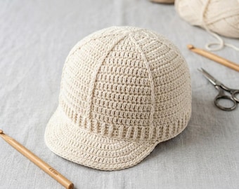 May include: A beige crochet baseball cap featuring a structured six-panel design with a curved brim. The hat is crafted from light-colored yarn and displays a consistent stitch pattern throughout the crown and the visor area.