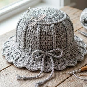 May include: A gray crocheted hat with a scalloped brim and a decorative silver butterfly. The hat features a bow and vertical ribbed detailing. The hat is handmade and sits on a wooden surface.