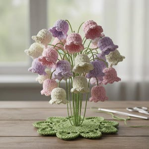 May include: A handcrafted crochet flower arrangement featuring bell-shaped flowers in pastel colors: pink, lavender, and cream. The flowers are attached to green wire stems, arranged in a green crochet base, and displayed on a wooden surface.