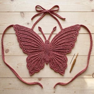 Puede incluir: Top de mariposa de ganchillo rosa polvoriento hecho a mano, con patrones de alas detallados y un lazo halter. La parte superior se muestra sobre una superficie de madera clara, con un ganchillo visible.