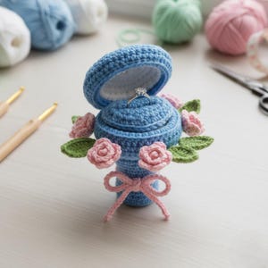 Puede incluir: Una caja de anillos azul tejida a ganchillo, abierta para revelar un anillo de diamantes. La caja está decorada con rosas rosas tejidas a ganchillo y hojas verdes, atadas con un lazo rosa. Hilo y ganchillos en el fondo.