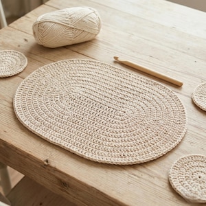 May include: A cream-colored, oval crochet placemat with matching coasters and a ball of yarn. A wooden crochet hook rests nearby on a wooden table. The handmade items are displayed on a light-colored wooden surface.