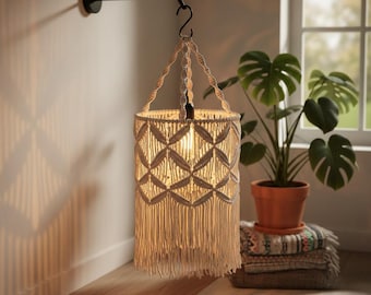 May include: A cream-colored macrame pendant light fixture with a geometric pattern and fringe. The light hangs from a black metal arm attached to a wall. A lit bulb is visible inside the shade. The fixture is a decorative home decor item.