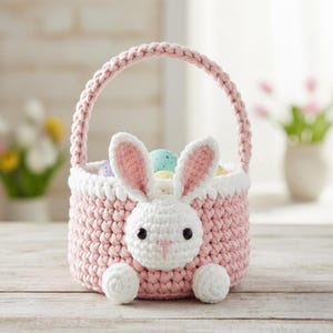 May include: A handmade pink crochet Easter basket with a white bunny face and ears. The basket has a handle and is filled with colorful Easter eggs. The bunny face has black eyes and a pink nose. The basket is sitting on a wooden surface.