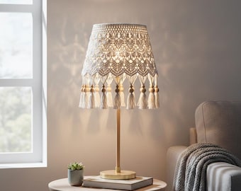May include: A cream-colored table lamp with a crocheted shade. The shade has a geometric pattern and is adorned with tassels and wooden beads. The lamp has a wooden base and stem, and sits on a round table with a small potted plant and a book.