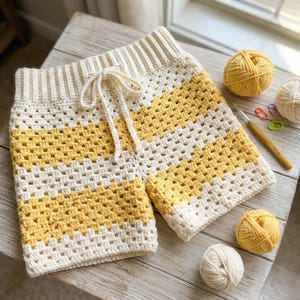 May include: Handmade crochet shorts in a granny square pattern. The shorts feature alternating stripes of cream and yellow. A cream-colored drawstring is visible at the waist. Yarn balls and a crochet hook are also in the image.