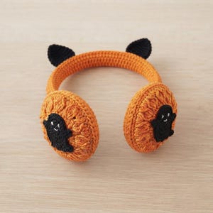 May include: Orange crochet earmuffs featuring a textured circular design with a small black ghost appliqué on each side and two pointed black cat ears attached to the top of the headband, creating a festive accessory for the autumn season.
