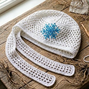 May include: A white crocheted hat with a blue snowflake applique and long, openwork ties. The hat is made of a light, airy yarn and is displayed on a rustic wooden surface. A crochet hook and other craft supplies are visible.