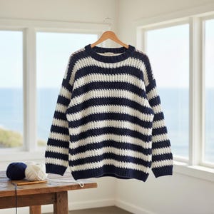 May include: A navy blue and white striped sweater hangs on a wooden hanger. The chunky knit sweater has a crew neck and long sleeves. Two balls of yarn and knitting needles sit on a wooden table in front of a window overlooking the ocean.