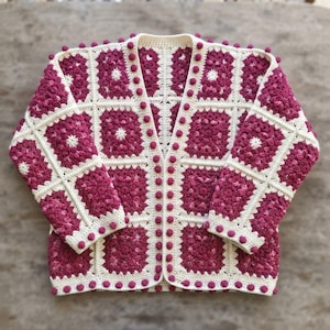 May include: A long-sleeve cardigan constructed from cream and magenta granny squares. The garment features a V-neckline and is finished with a decorative border of small, raised magenta bobbles along the front opening, cuffs, and hemline.