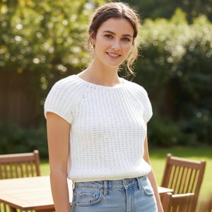 Könnte beinhalten: Ein weißes, kurzärmeliges Häkeltop mit rundem Ausschnitt. Das Oberteil hat ein strukturiertes, geripptes Design und wird mit hellblauen Jeans getragen. Das Bild ist im Freien mit einem verschwommenen grünen Hintergrund aufgenommen.
