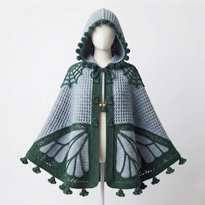 May include: A light blue and dark green hooded crochet cape featuring a waffle stitch pattern. The garment includes dark green spiderweb motifs at the shoulders, butterfly wing patterns at the hem, and small ghost-shaped toggles with wooden beads.
