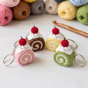 Puede incluir: Cuatro llaveros de gajos de pastel de ganchillo en rosa, marrón, amarillo y verde, cada uno coronado con glaseado blanco y una cereza roja. Los llaveros están en anillos plateados, con hilo y un ganchillo en el fondo.