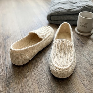 Könnte beinhalten: Ein Paar cremefarbene, gehäkelte Loafer. Die Schuhe haben ein klassisches Slip-on-Design mit gerippter Textur auf der Oberseite. Eine graue Decke und eine Tasse mit "COZY VIBES" befinden sich im Hintergrund. Die Schuhe stehen auf einer Holzoberfläche.