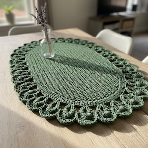 Peut inclure: Nappe ovale en crochet vert faite à la main. Le chemin de table présente un motif de point texturé et un bord festonné. Un petit vase en verre avec des brins de lavande est posé sur la table en bois clair.
