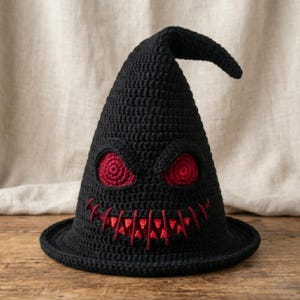May include: A black crochet witch hat with a pointed top and a wide brim. The hat features red eyes and a stitched red mouth, creating a spooky face design. The hat is displayed on a wooden surface.