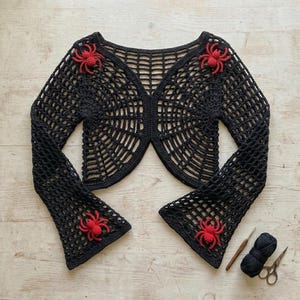 Puede incluir: Bolero negro de ganchillo con diseño de telaraña y mangas acampanadas. El bolero está adornado con tres arañas rojas de ganchillo. También se ven un ganchillo, hilo negro y tijeras.