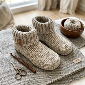May include: Two beige crochet slipper boots with ribbed cuffs, presented on a grey blanket. A wooden crochet hook and gold scissors are next to the slippers. A wooden bowl with yarn and a "Handmade with Love" tag are also visible.