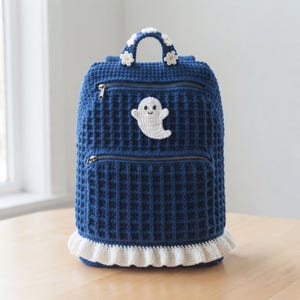 Könnte beinhalten: Ein dunkelblauer Häkelrucksack mit Waffelstruktur, verziert mit einer weißen Geister-Applikation auf der Vorderseite, einem weißen Rüschensaum an der Basis und einem Tragegriff, der mit kleinen weißen Häkelblumen geschmückt ist. Zwei Reißverschlussfächer.