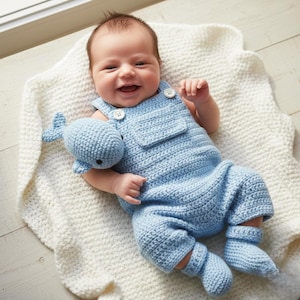 May include: A baby wearing a light blue crochet outfit with a matching whale toy. The outfit includes overalls and booties. The baby is lying on a white, textured blanket.