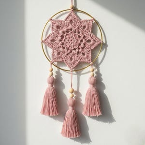May include: A handmade, pink crochet dream catcher with a gold metal ring. The dream catcher features a star-shaped crochet design in the center, with three tassels and wooden beads hanging below. The tassels and crochet are a soft pink color.