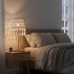 Op de afbeelding: Een beige tafellamp met een patroon lampenkap en kwastjes staat op een houten nachtkastje. De lamp verlicht een slaapkamer met een bed, kussens en een kleine potplant. De kamer wordt verlicht door zacht, warm licht.