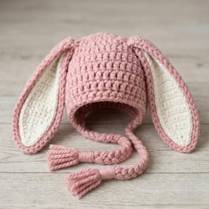 May include: A handmade pink crochet bunny hat with long, floppy ears. The inside of the ears is white. The hat has braided ties with tassels. The hat is designed for babies and toddlers.