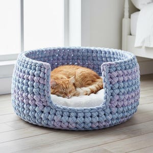 May include: A round, blue and purple crocheted cat bed with a white interior cushion. An orange tabby cat is curled up asleep inside the bed. The bed is made of thick, soft yarn and sits on a light wood floor.