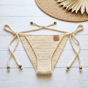 Puede incluir: Braguita de bikini de ganchillo color crema con laterales anudados y detalles de cuentas de madera. La braguita de bikini tiene una pequeña etiqueta rectangular con el texto "HHC" y "100% Cotton". La braguita de bikini se muestra sobre una superficie de madera blanca.