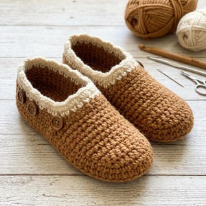 May include: A pair of handmade brown crochet slippers with a cream-colored trim and wooden buttons. The slippers are displayed on a light wooden surface, with yarn and crochet tools in the background. The slippers are a cozy, handcrafted item.