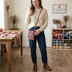 May include: A woman wearing a cream-colored sweater and blue jeans, holding a small, reddish-pink crossbody bag. The bag has a flap closure and a long strap. The background includes a wooden table, a shelf with yarn, and a sign that says "The Crochet Corner - Handmade Goods."
