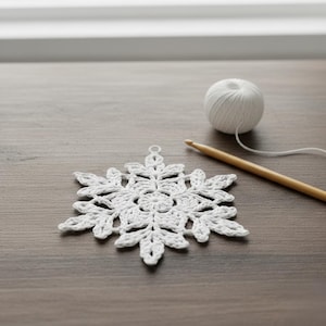 Puede incluir: Adorno de copo de nieve blanco tejido a ganchillo con un diseño floral detallado. Una madeja de hilo blanco y un ganchillo de madera están junto al copo de nieve sobre una superficie de madera. Decoración navideña de invierno.