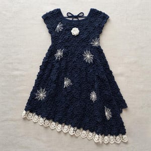 May include: A dark navy blue crochet dress featuring a textured floral pattern, short sleeves, and a tie-back neckline. The dress is decorated with white embroidered spider web motifs and finished with a white lace trim featuring a skull pattern.