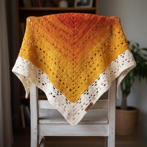 Puede incluir: Un chal de ganchillo hecho a mano en un degradado de naranja, amarillo y crema. El chal está drapeado sobre una silla de madera blanca. El diseño presenta un patrón calado y un borde color crema.